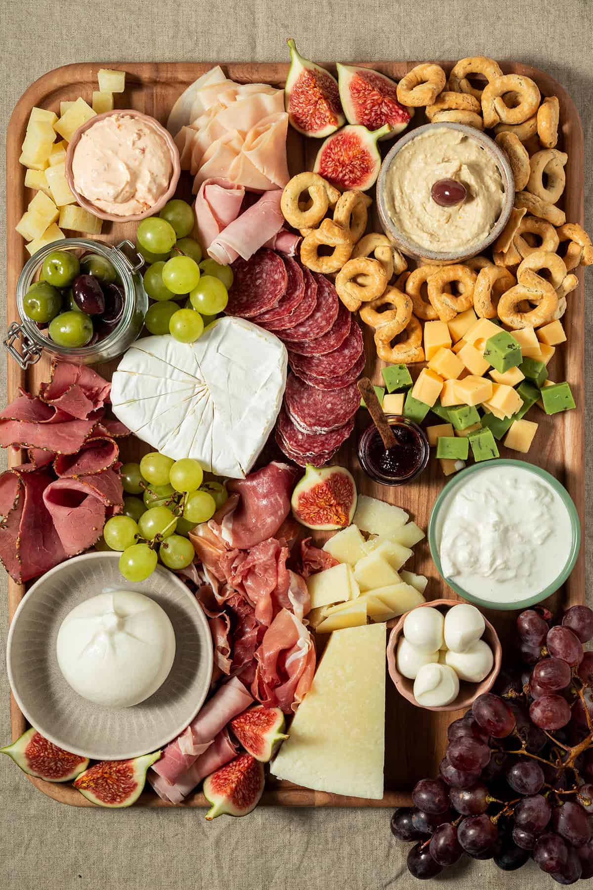 Vollst&auml;ndig angerichtete Charcuterie-Platte mit K&auml;se, Wurst, Feigen, Trauben, Crackern, Dips und Antipasti auf einem Holzbrett.