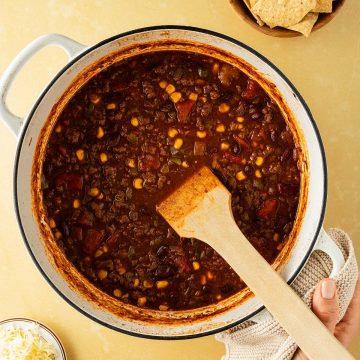 Ein Hand h&auml;lt einen gro&szlig;en Topf mit Chili con Carne, Nachos und K&auml;se im Bildrand.