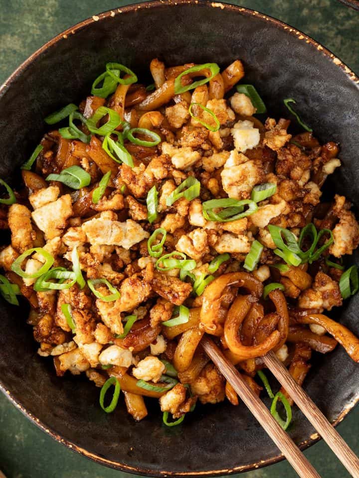 Eine Sch&uuml;ssel Udon Nudeln mit knusprigen Tofu-Hack und Fr&uuml;hlingszwiebeln auf dunkelgr&uuml;nem Untergrund.