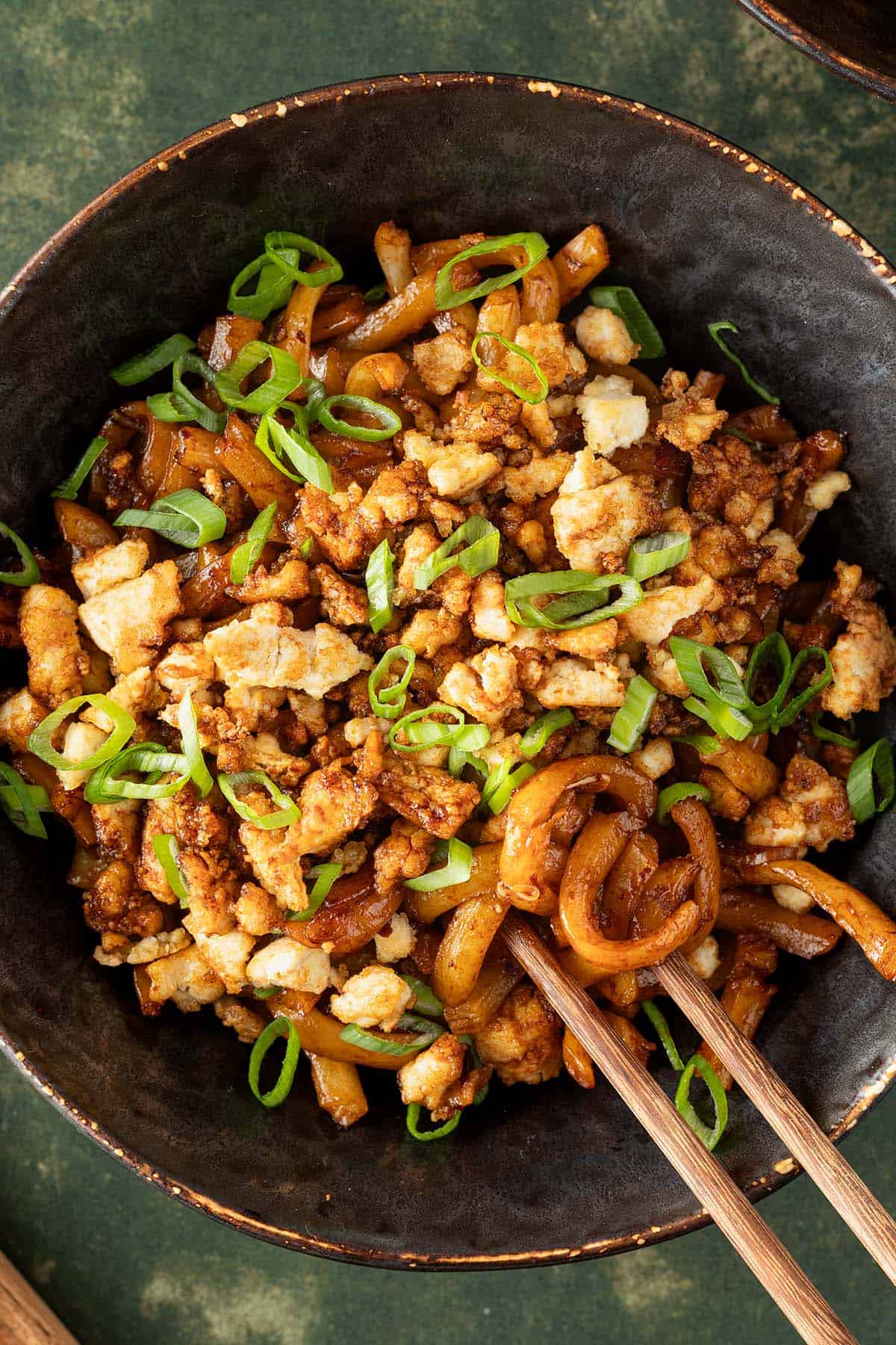 Eine Sch&uuml;ssel Udon Nudeln mit knusprigen Tofu-Hack und Fr&uuml;hlingszwiebeln auf dunkelgr&uuml;nem Untergrund.