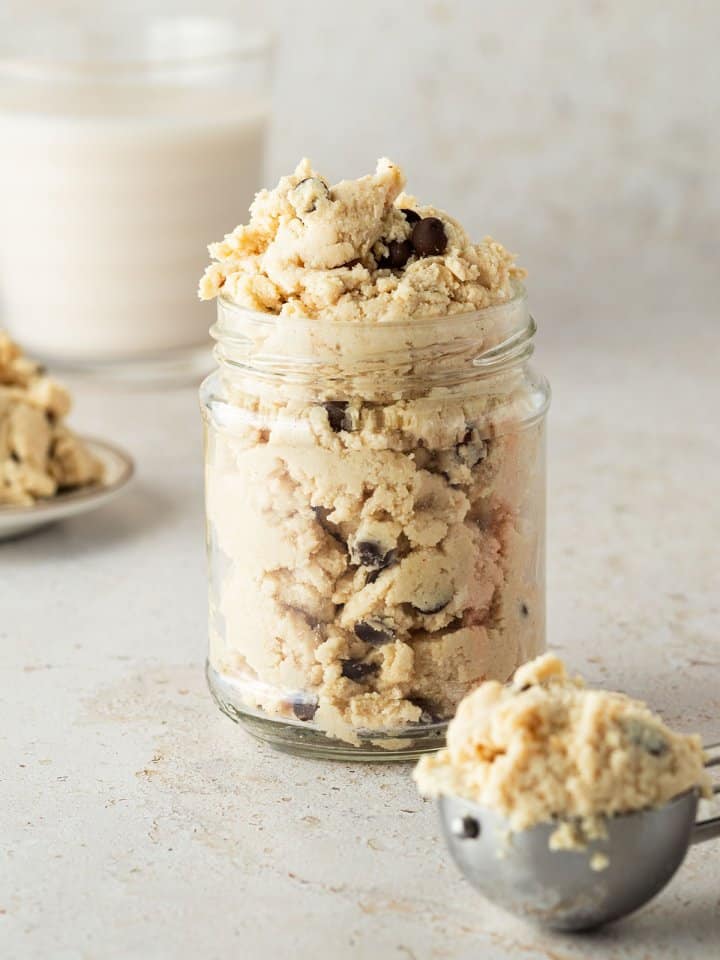 Ein Glas essbarer Cookie Dough vor beigen Hintergrund, davor ein Eisportionierer mit einer Portion Cookie Dough.