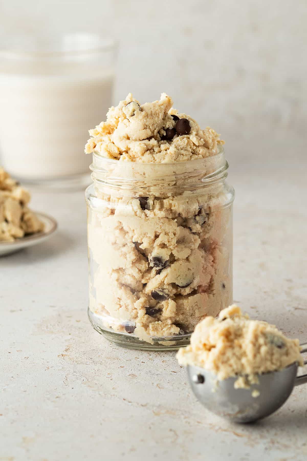 Ein Glas essbarer Cookie Dough vor beigen Hintergrund, davor ein Eisportionierer mit einer Portion Cookie Dough.