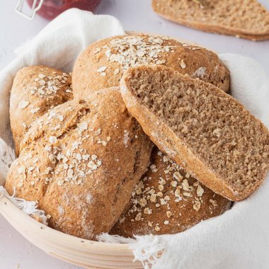 Ein Brotkorb mit Vollkornbrötchen auf einem weißen Untergrund, im Hintergrund ein aufgeschnittenes Brötchen und ein Glas Marmelade.