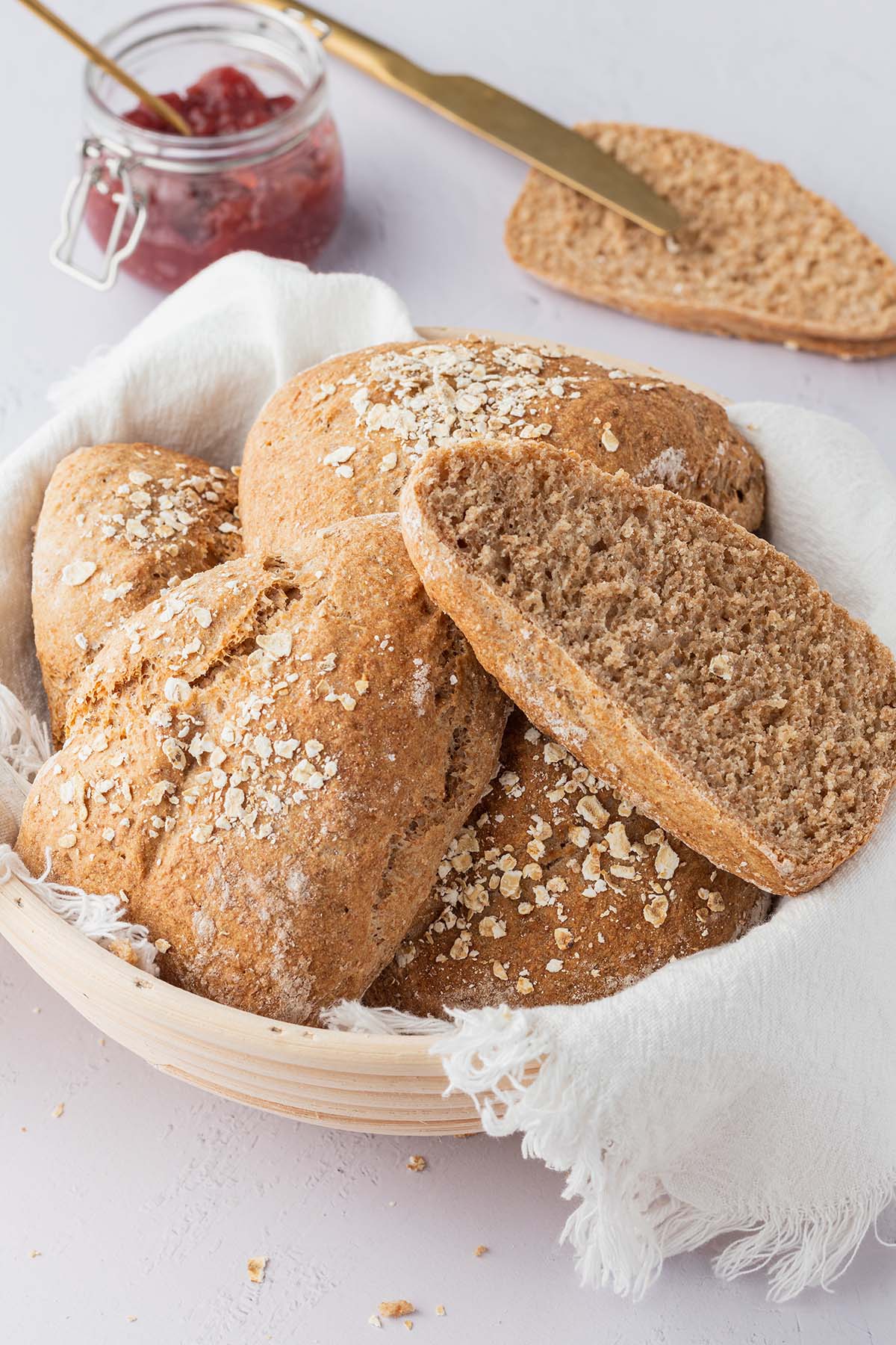 Ein Brotkorb mit Vollkornbr&ouml;tchen auf einem wei&szlig;en Untergrund, im Hintergrund ein aufgeschnittenes Br&ouml;tchen und ein Glas Marmelade.