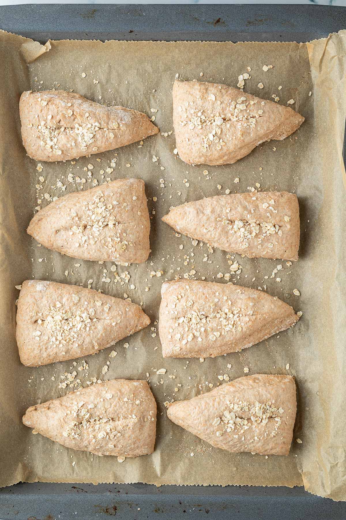 Acht zu Dreiecken geformten und mit Haferflocken bestreuten Brötchen vor dem Backen auf einem Blech.