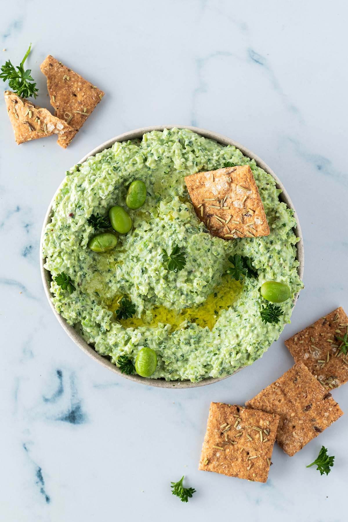 Edamame Dip in einer Sch&uuml;ssel, serviert mit Crackern und frischer Petersilie.