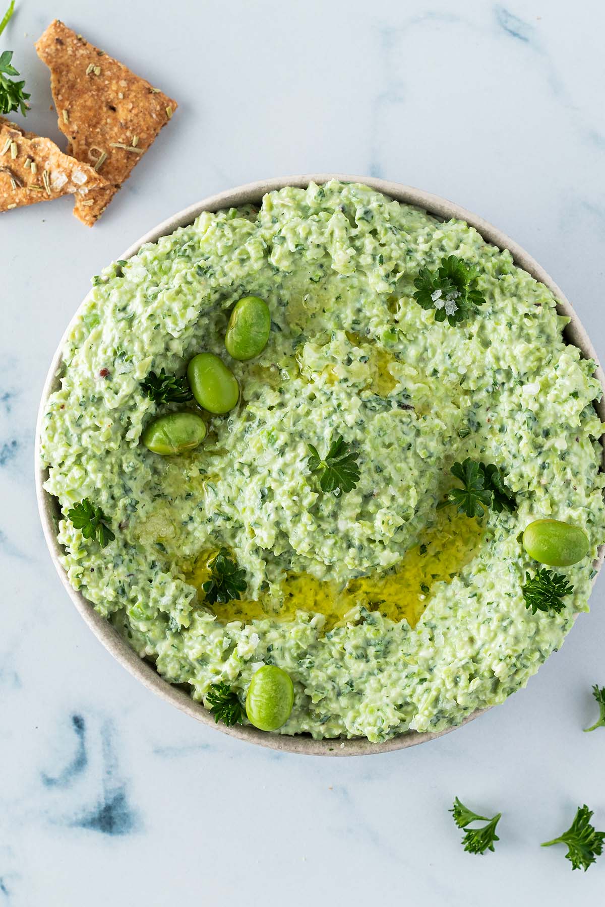 Vogelperspektive auf einen Edamame Dip, mit Cracker und Petersilie im Bildrand.