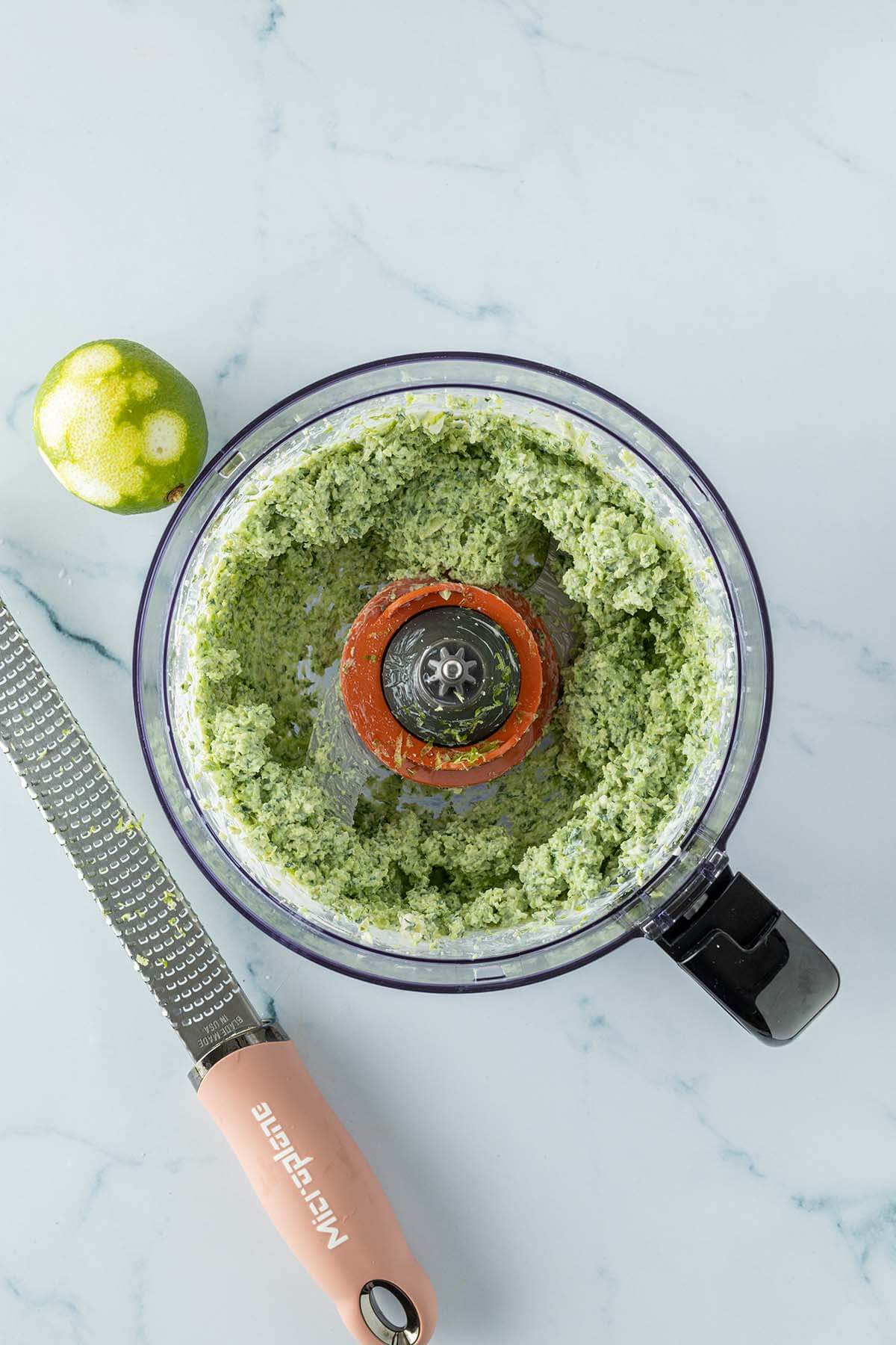 Vogelperspektive auf einen Food Processor, in dem sich der Edamame Dip befindet, daneben eine Reibe und eine Limette, wo ein Teil der Schale abgerieben wurde.