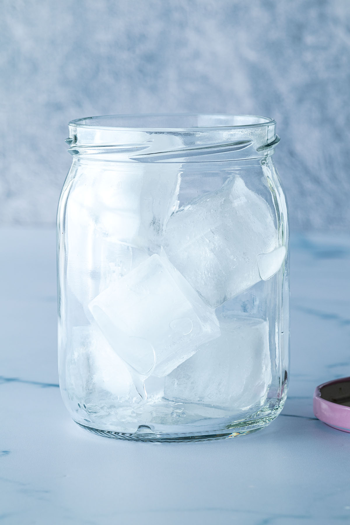 Ein mit Eisw&uuml;rfeln gef&uuml;lltes Schraubglas vor blau-grauen Hintergrund.