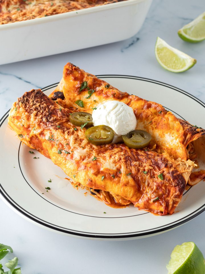 Zwei mit K&auml;se &uuml;berbackene Enchiladas auf einem Teller mit Sourcream und Jalape&ntilde;os serviert, Deko und eine Auflaufform mit weiteren Enchiladas im Bildrand.