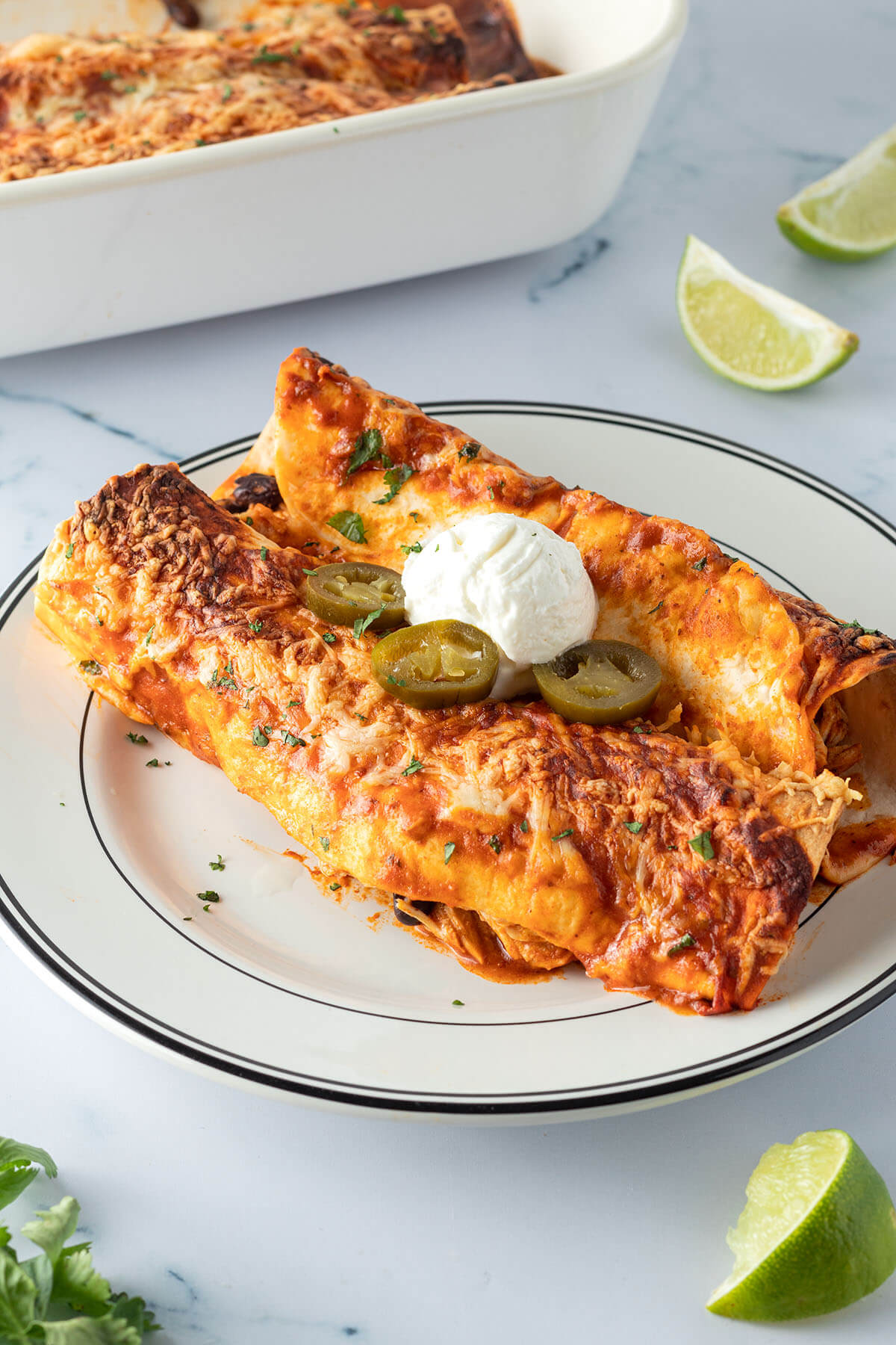 Zwei mit K&auml;se &uuml;berbackene Enchiladas auf einem Teller mit Sourcream und Jalape&ntilde;os serviert, Deko und eine Auflaufform mit weiteren Enchiladas im Bildrand.