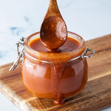 L&ouml;ffel wird in ein Glas mit einer dickfl&uuml;ssigen Enchilada-Sauce getaucht, das auf einem Holzbrett steht.