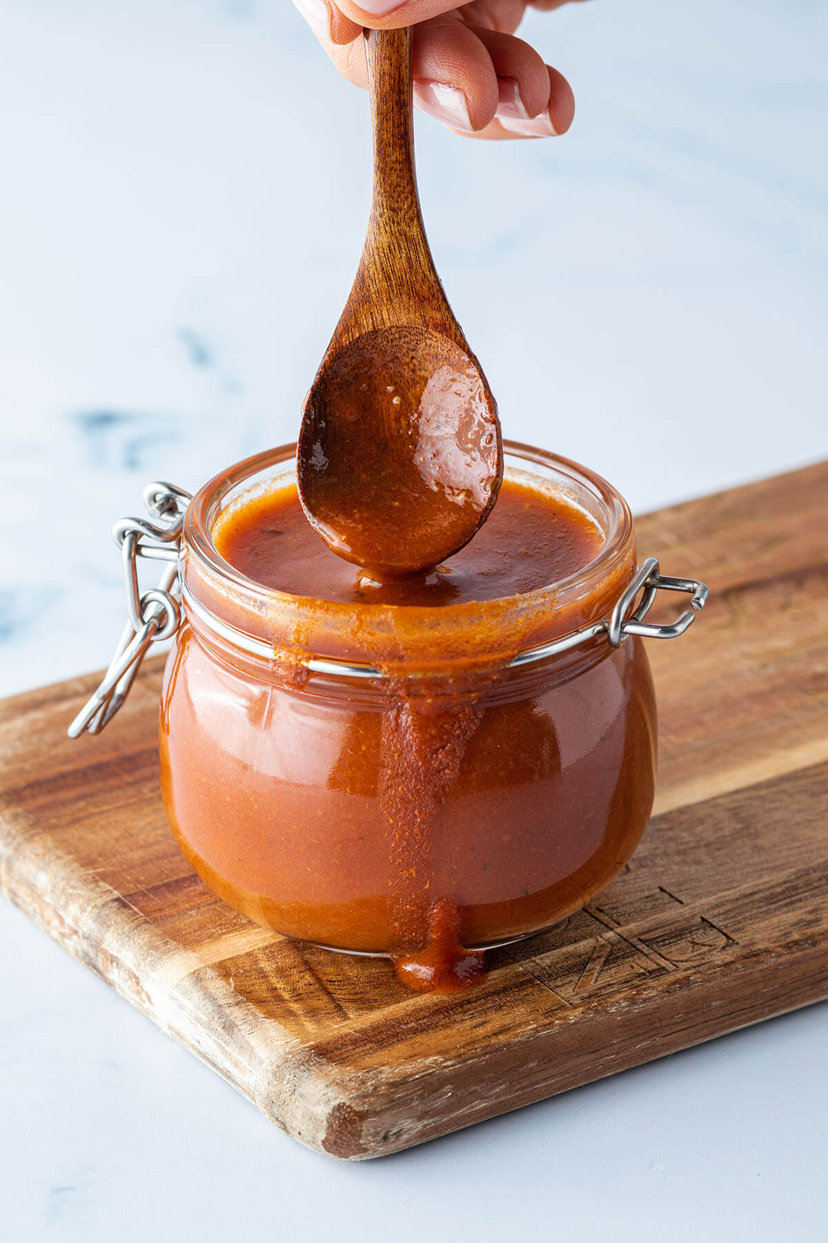 L&ouml;ffel wird in ein Glas mit einer dickfl&uuml;ssigen Enchilada-Sauce getaucht, das auf einem Holzbrett steht.