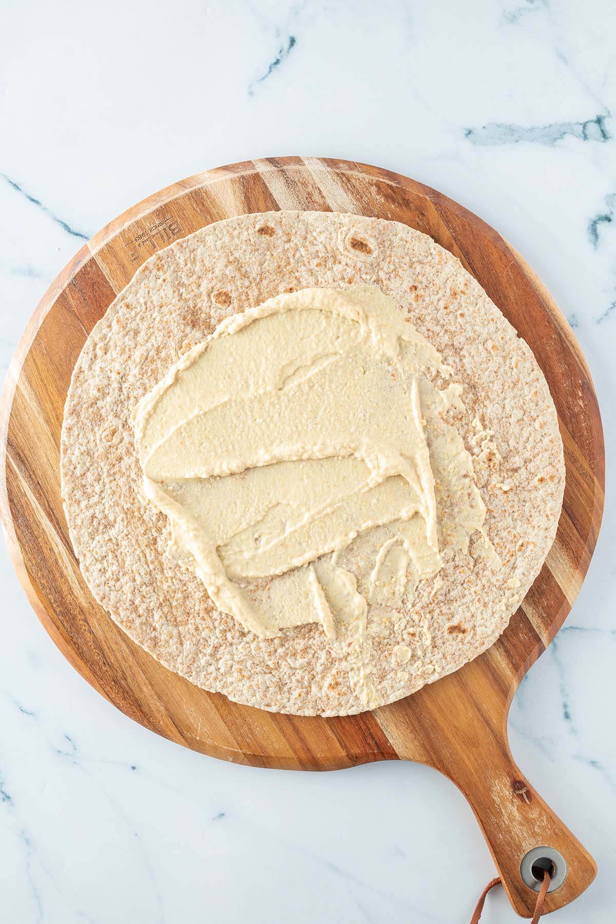 Hummus auf einem Vollkorn-Wrap, der auf einem runden Holzbrett liegt.