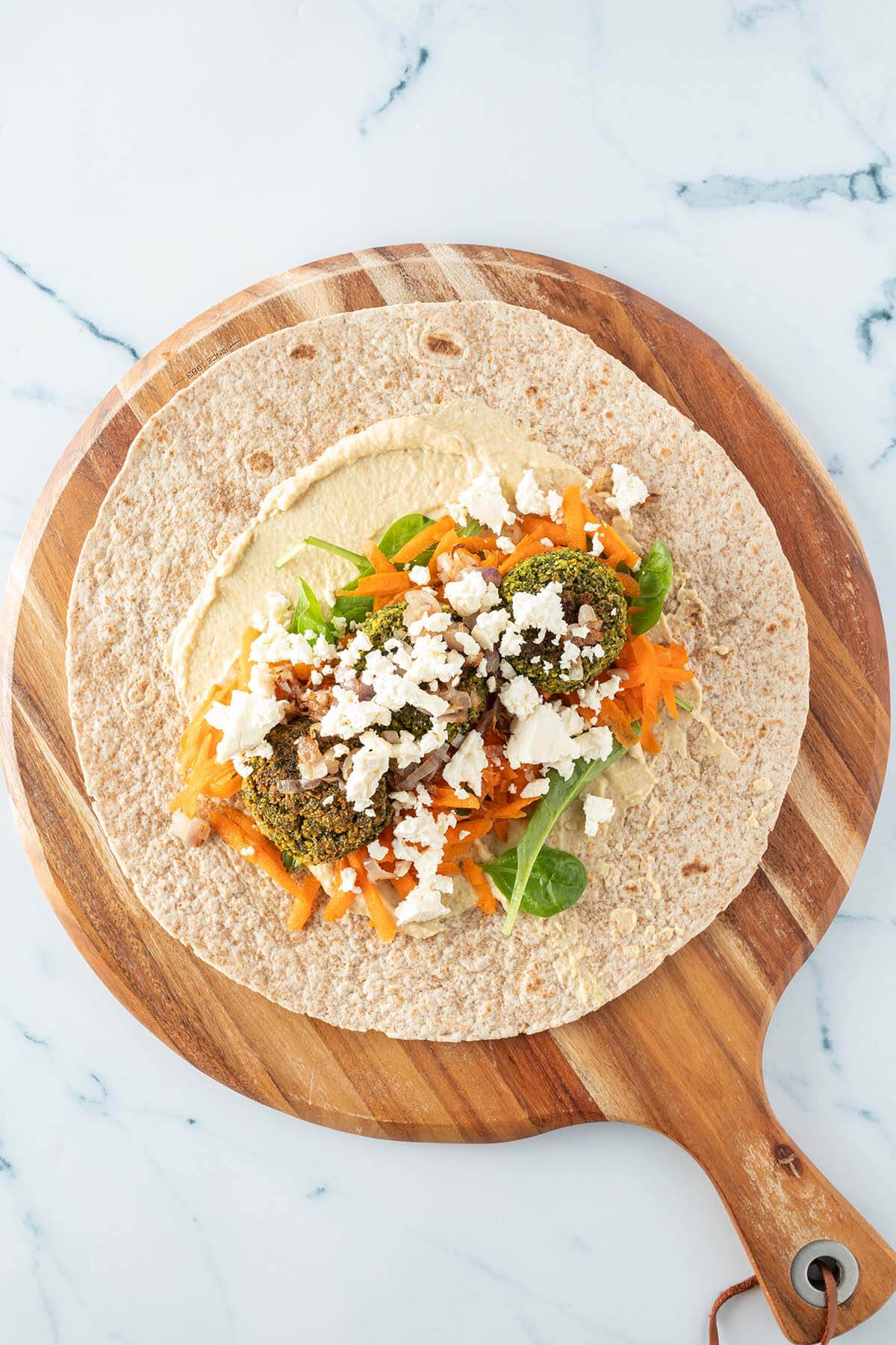 Wrap mit Hummus, Gem&uuml;se, Falafel und Feta belegt auf einem Holzbrett.