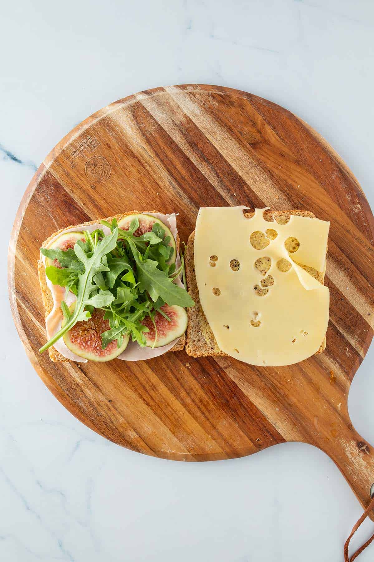Eine mit K&auml;se und eine mit Schinken, frischen Feigen und Rucola belegte Toastscheibe nebeneinander auf einem Brett.