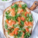 Knuspriger Flammkuchen mit Lachs, Rucola und Pesto auf einem Holzbrett serviert.