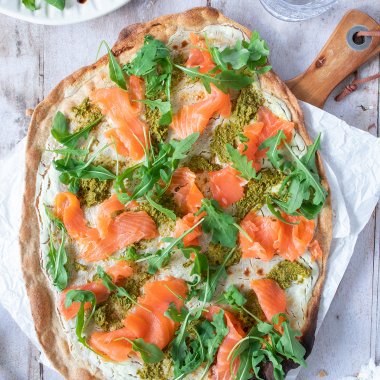 Knuspriger Flammkuchen mit Lachs, Rucola und Pesto auf einem Holzbrett serviert.