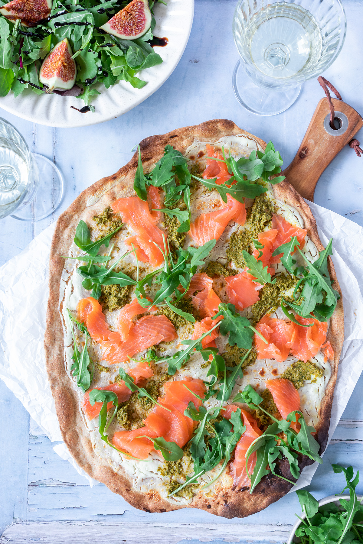 Knuspriger Flammkuchen belegt mit Lachs, Rucola und gr&uuml;nem Pesto, serviert mit einem Salat und Wei&szlig;wein.