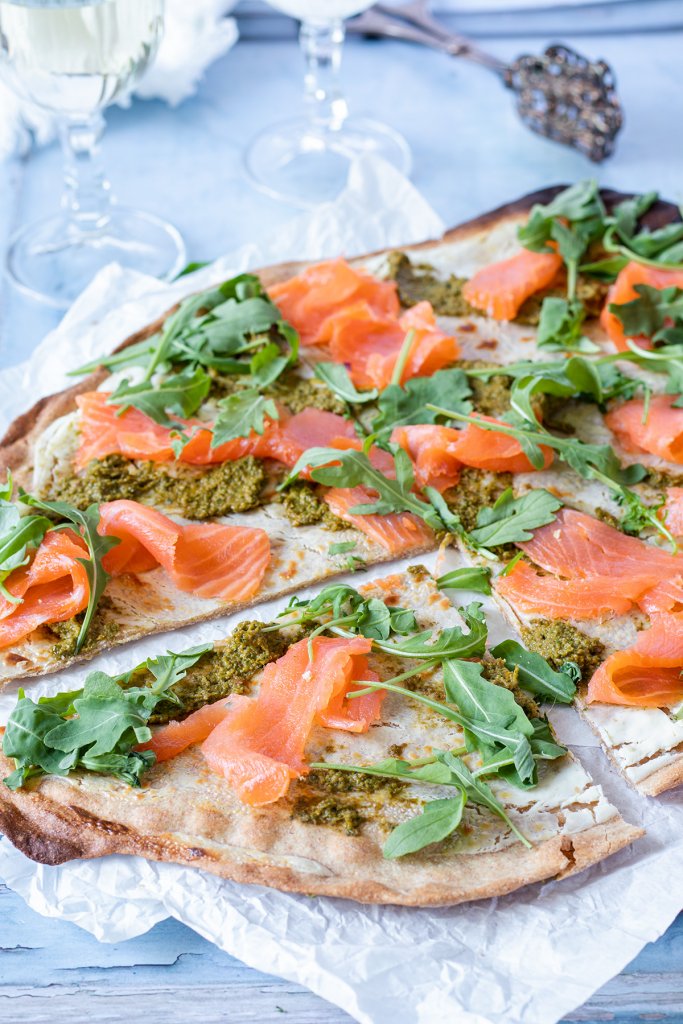 Flammkuchen mit Lachs, Rucola und Pesto belegt, im Hintergrund Deko.