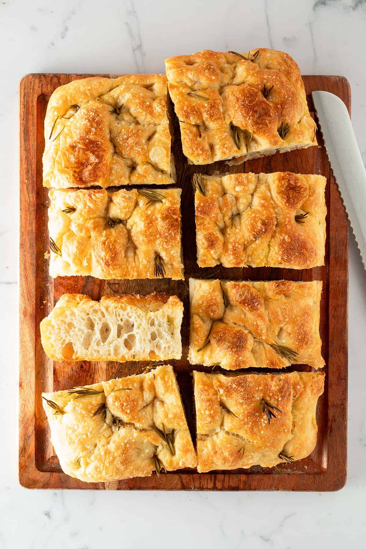 In rechteckige St&uuml;cke geschnittene Focaccia mit Rosmarin und grobem Salz auf einem Holzbrett, daneben ein Brotmesser.