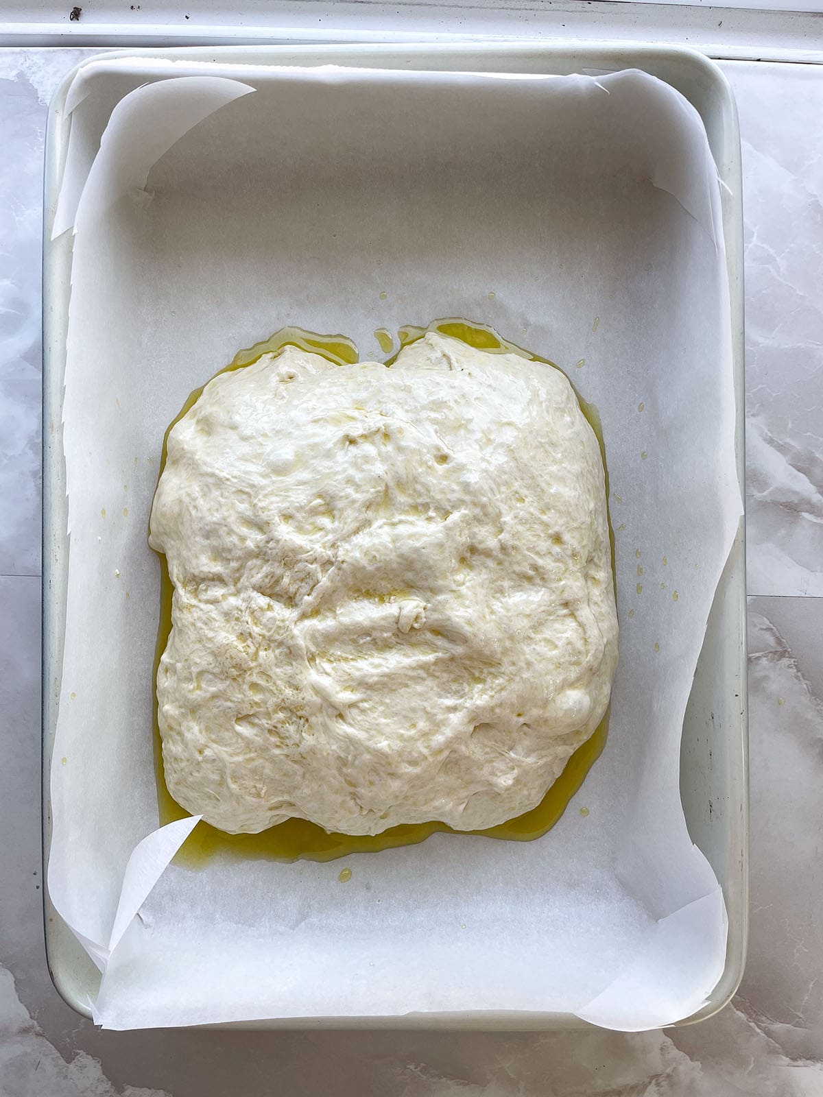 Focaccia-Teig auf Oliven&ouml;l in einer mit Backpapier ausgelegten Backform vor der zweiten Gehzeit.