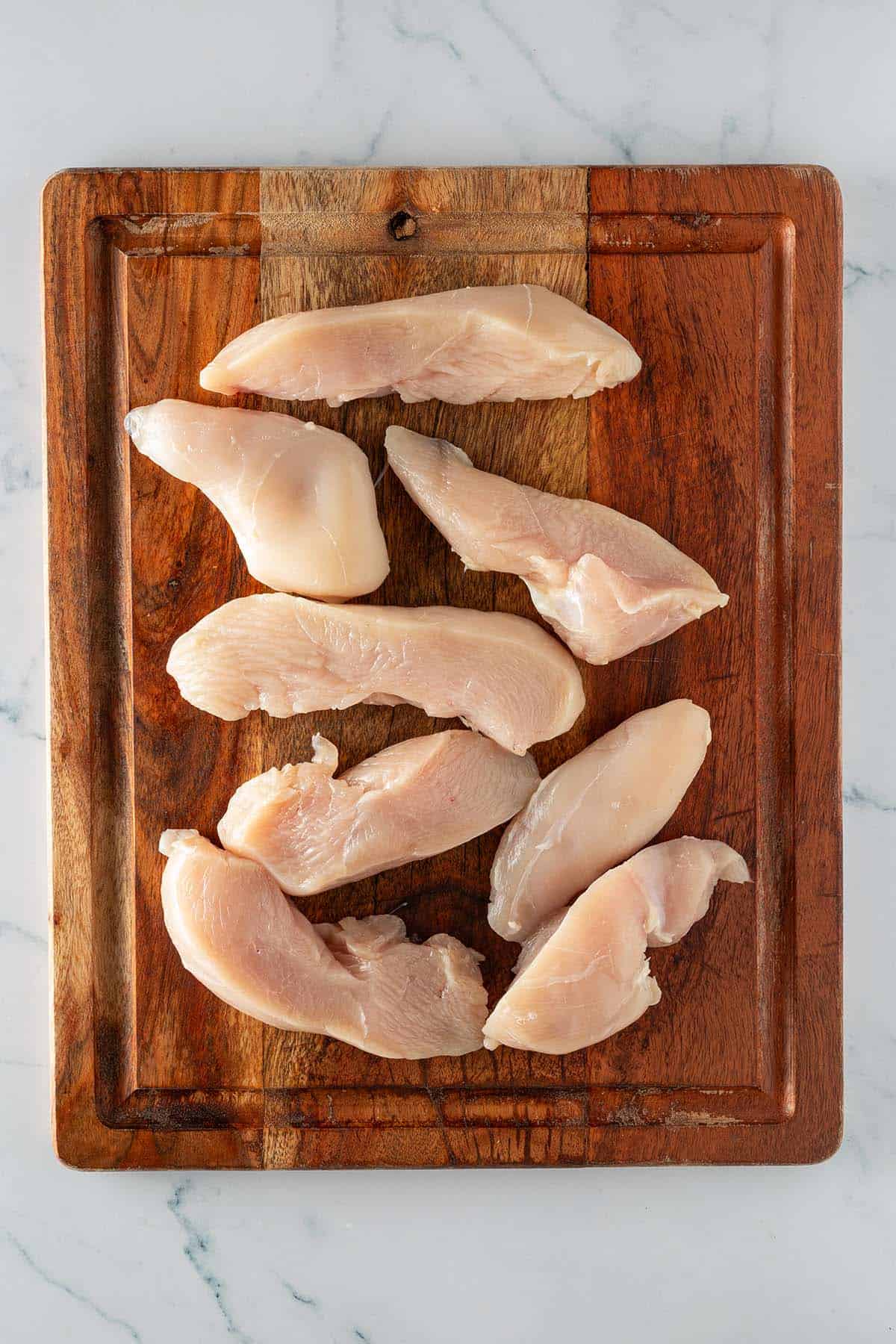Rohe in circa 4 Zentimeter dicke Streifen geschnittene H&auml;hnchenbrustfilets auf einem Holzbrett.