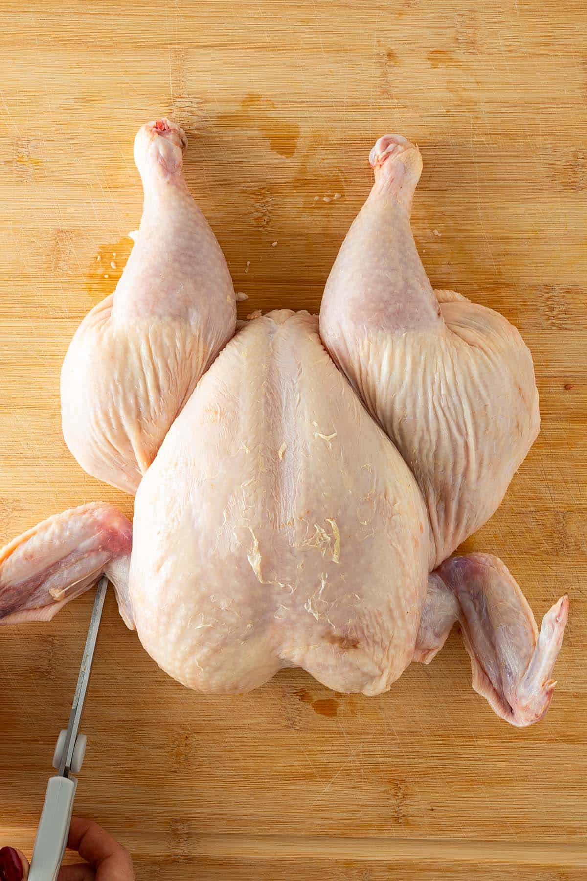 Ganze rohes H&auml;hnchen auf einem Holzbrett, die Haut an den Fl&uuml;geln wird mit einer Schere eingeschnitten.