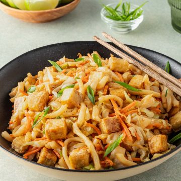 Eine Schüssel gebratene Udon Nudeln mit Tofu und Gemüse, Deko im Hintergrund.