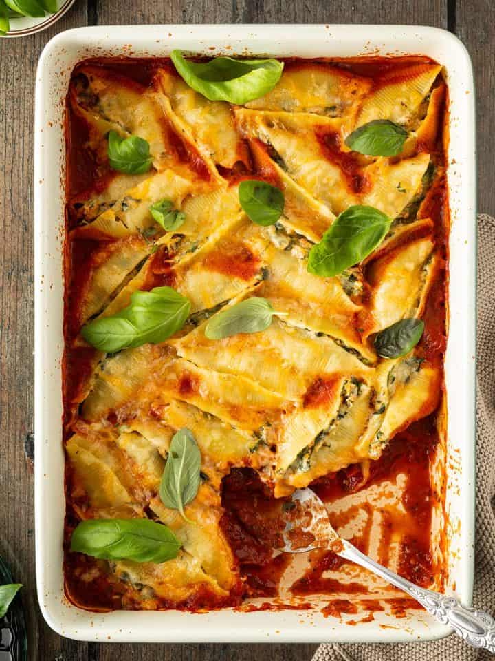 Eine Auflaufform mit gef&uuml;llten Muschelnudeln in Tomatensauce und mit frischem Basilikum, wo mit einem L&ouml;ffel bereits eine Portion herausgenommen wurde.