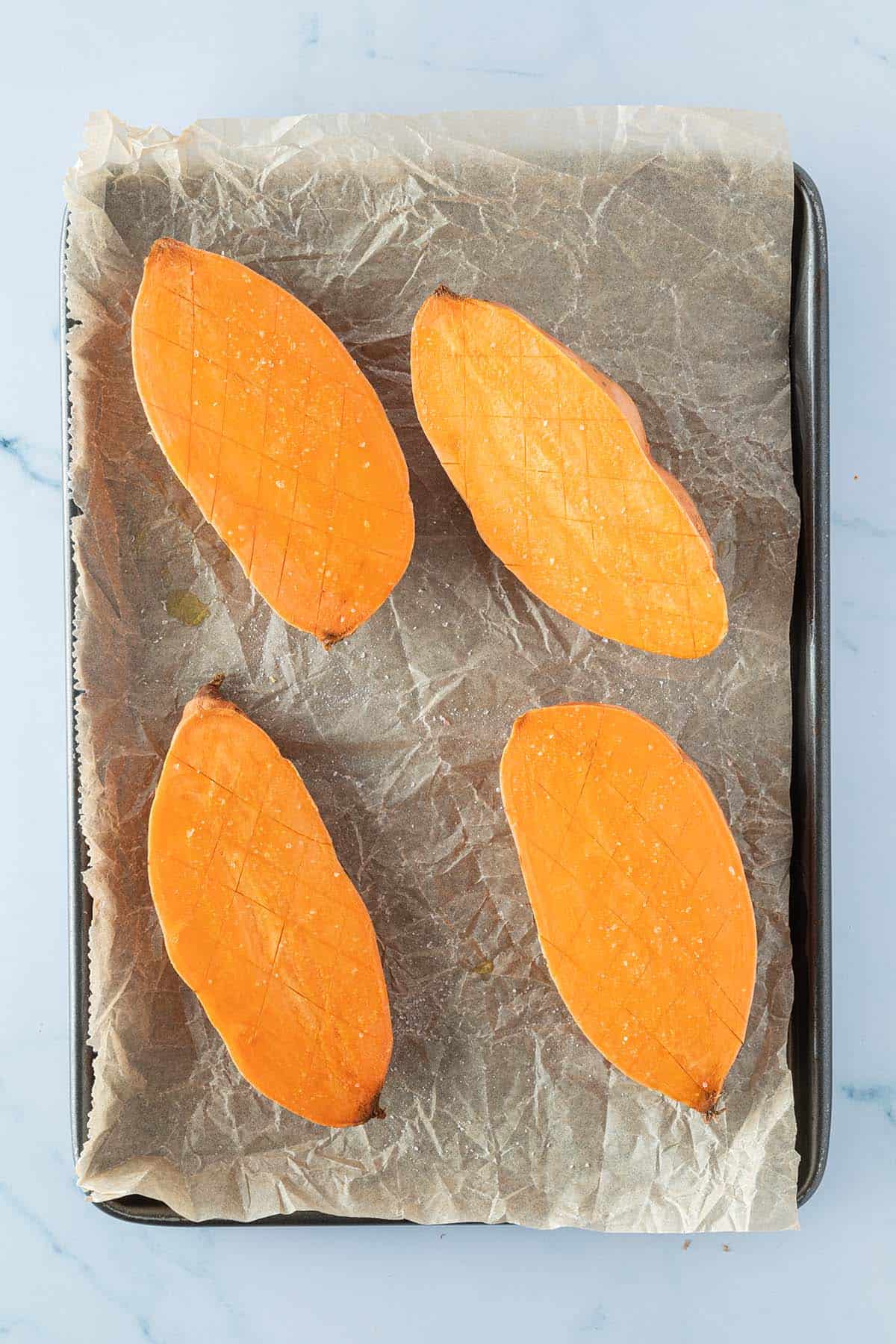 Zwei halbierte Süßkartoffeln, deren Schnittfläche rautenförmig eingeritzt und mit Öl und Salz eingerieben wurde, auf einem Backblech.