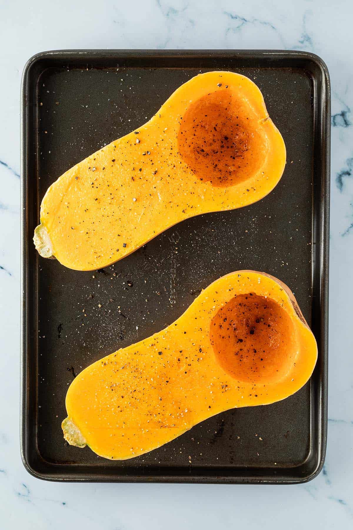 Zwei mit &Ouml;l, Salz und Pfeffer marinierte Butternut K&uuml;rbish&auml;lften auf einem Backblech.