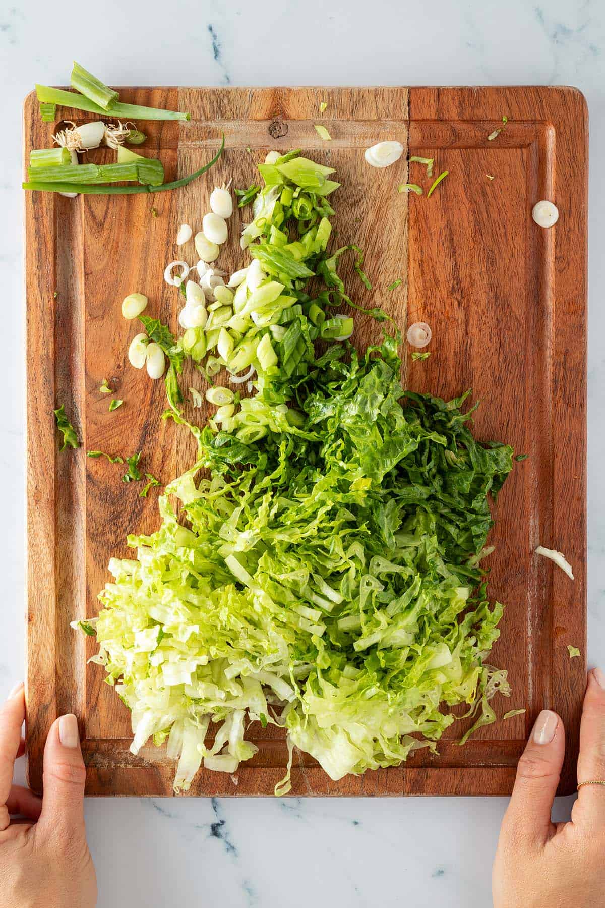 In feine Streifen geschnittener Salat und in d&uuml;nne Ringe geschnittene Fr&uuml;hlingszwiebeln auf einem Holzbrett, gehalten von zwei H&auml;nden.