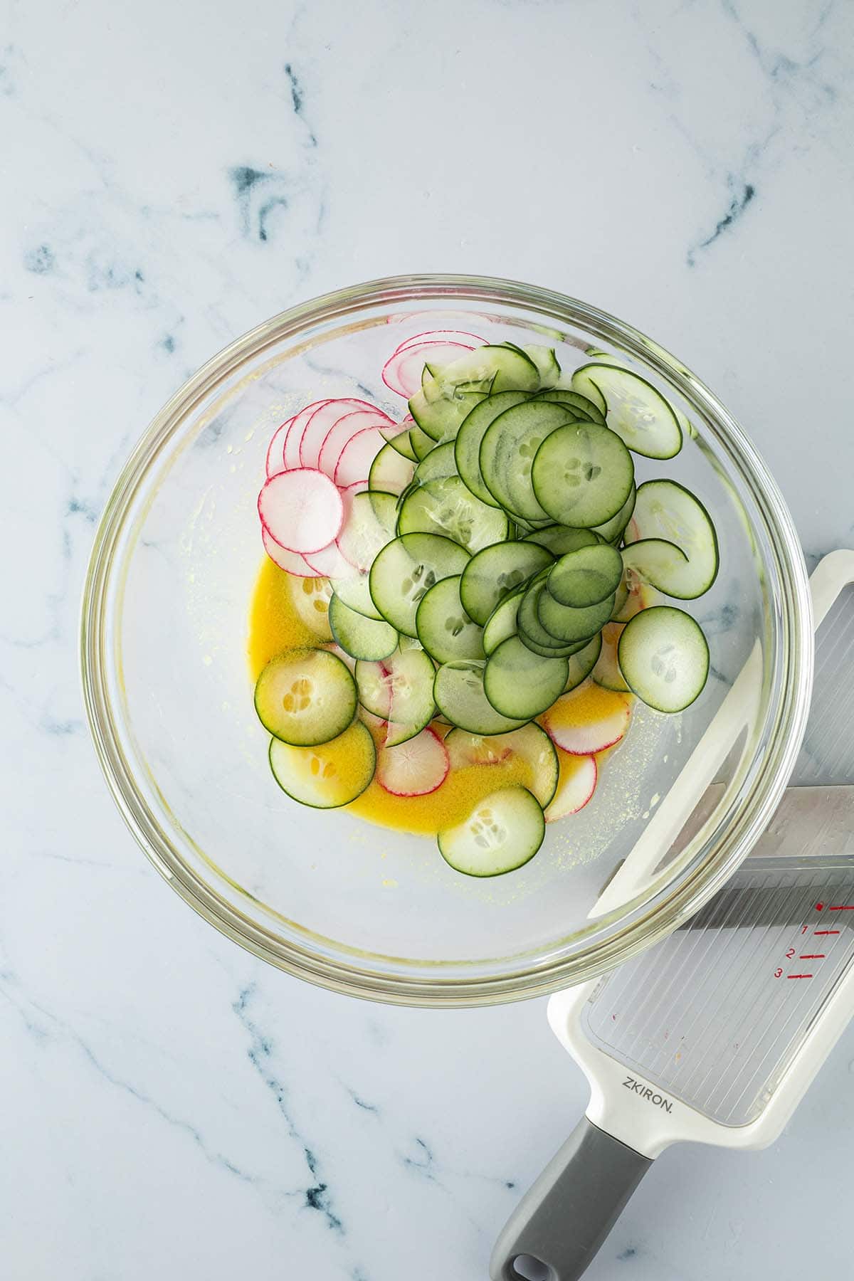 Eine Salatsch&uuml;ssel mit Dressing und in d&uuml;nne Scheibchen gehobelte Radieschen und Gurken, daneben eine Gem&uuml;sehobel.