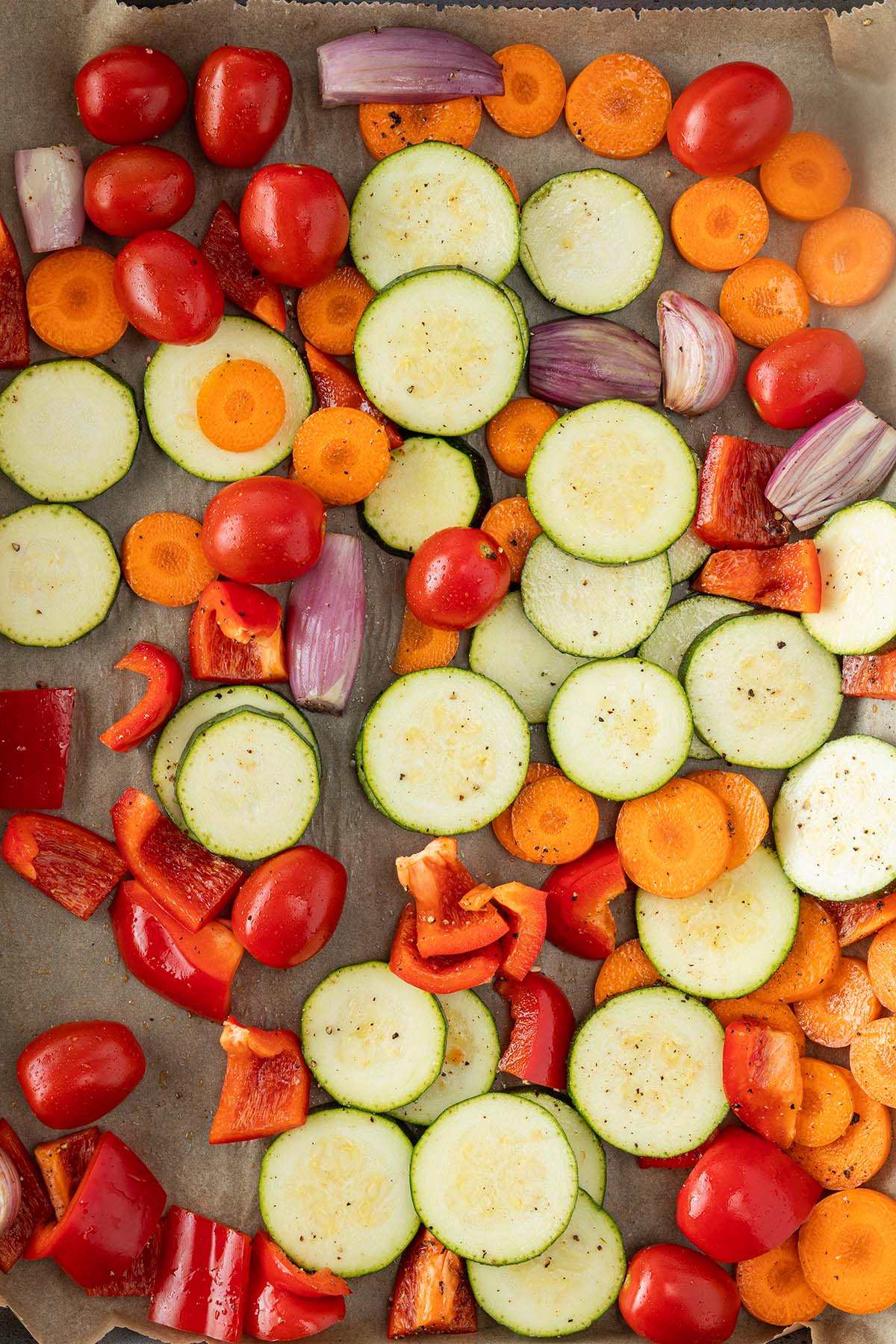 In Scheiben geschnittene Zucchini und Karotte liegen zusammen mit Paprikast&uuml;ckchen, Schalotten, Knoblauchzehen und Kirschtomaten auf einem Backblech.