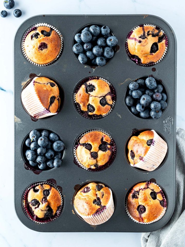 Vogelperspektive auf ein Muffinblech mit Blaubeer-Muffins und Blaubeeren, Deko im Bildrand.
