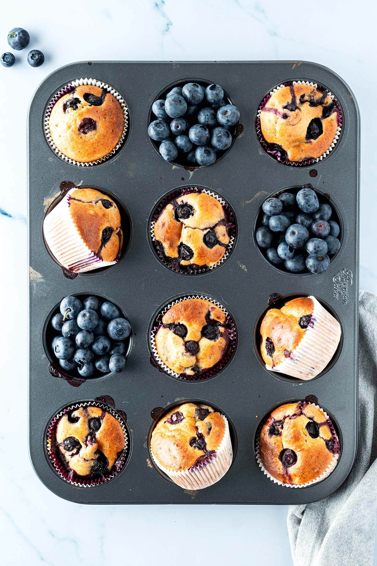 Vogelperspektive auf ein Muffinblech mit Blaubeer-Muffins und Blaubeeren, Deko im Bildrand.