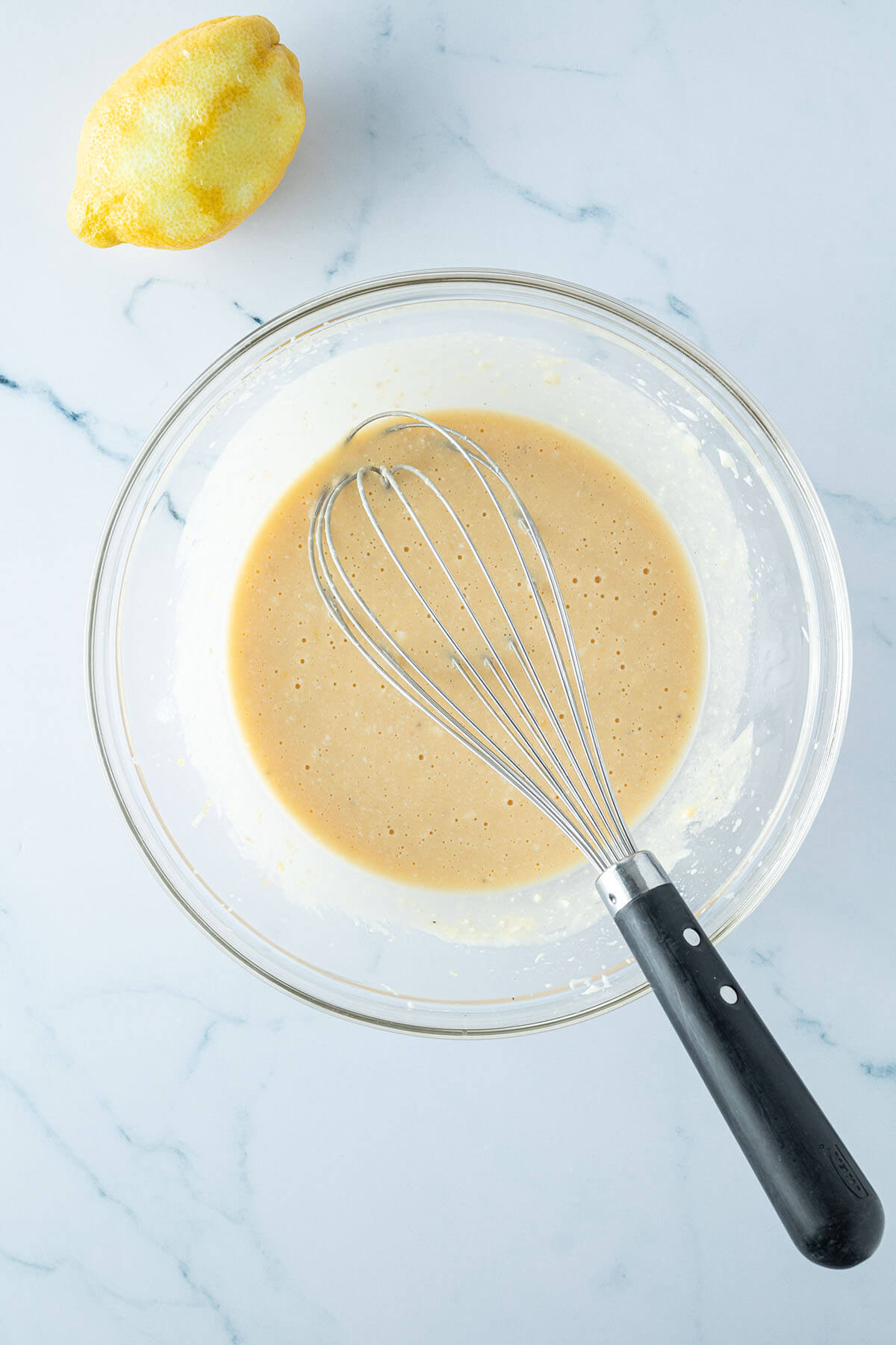Fl&uuml;ssige Zutaten f&uuml;r die Muffins in einer Glassch&uuml;ssel mit einem Schneebesen verr&uuml;hrt, im Bildrand eine abgeriebene Zitrone.