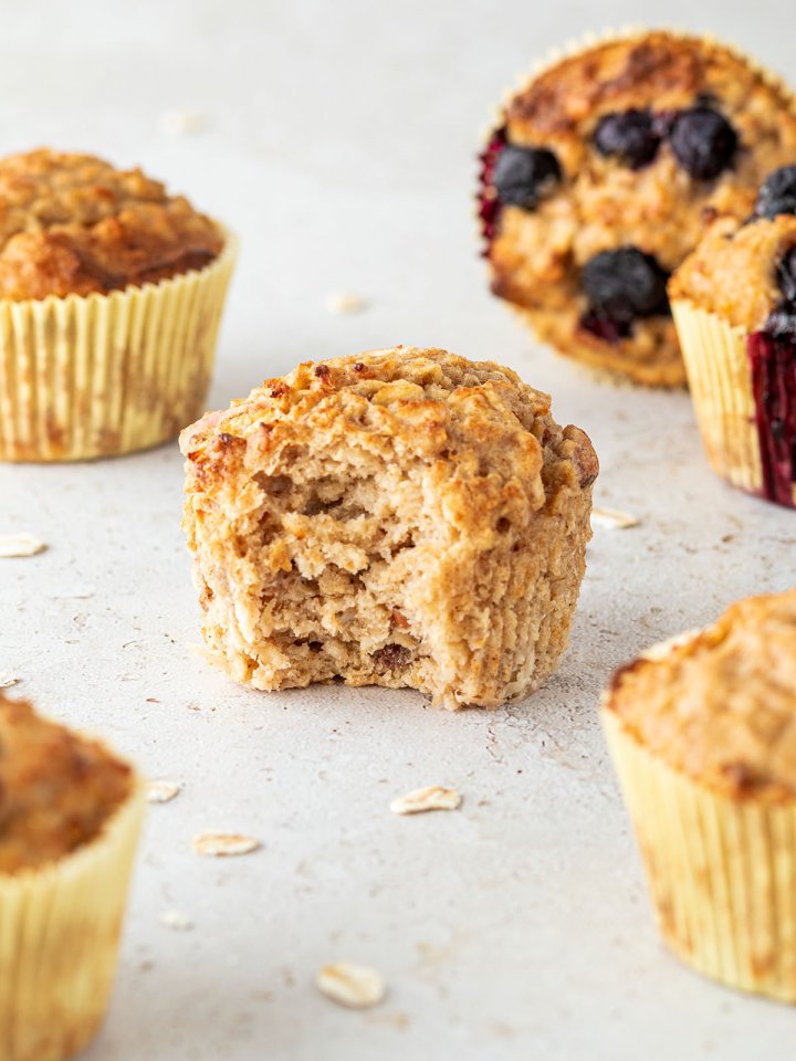 Ein angebissener Fr&uuml;hst&uuml;cksmuffin auf einem hellen Untergrund, daneben sind weitere Muffins und ein paar Haferflocken.