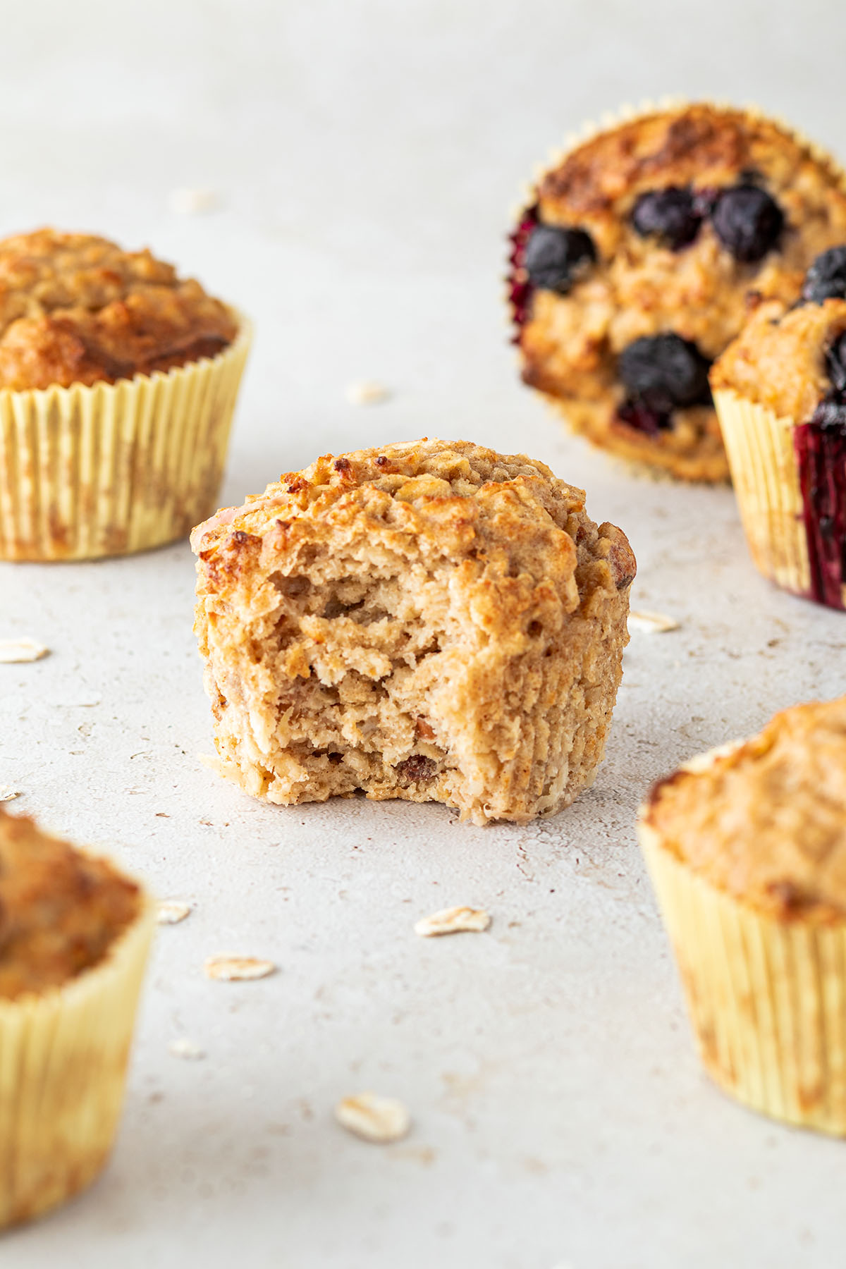 Ein angebissener Fr&uuml;hst&uuml;cksmuffin auf einem hellen Untergrund, daneben sind weitere Muffins und ein paar Haferflocken.