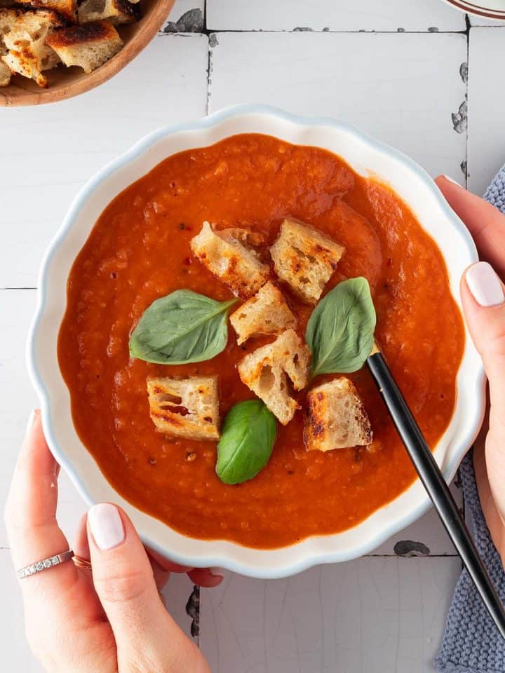 Zwei H&auml;nde halten eine Sch&uuml;ssel Tomatensuppe mit Croutons und Basilikum.