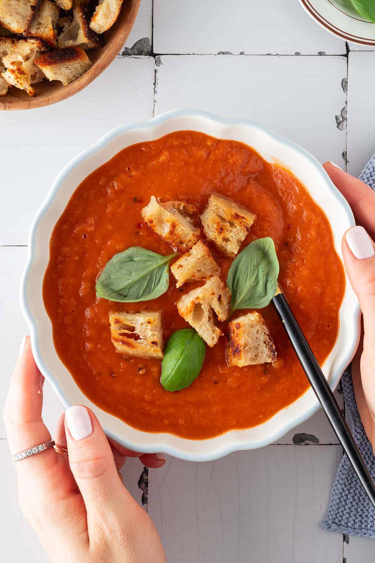 Zwei H&auml;nde halten eine Sch&uuml;ssel Tomatensuppe mit Croutons und Basilikum.