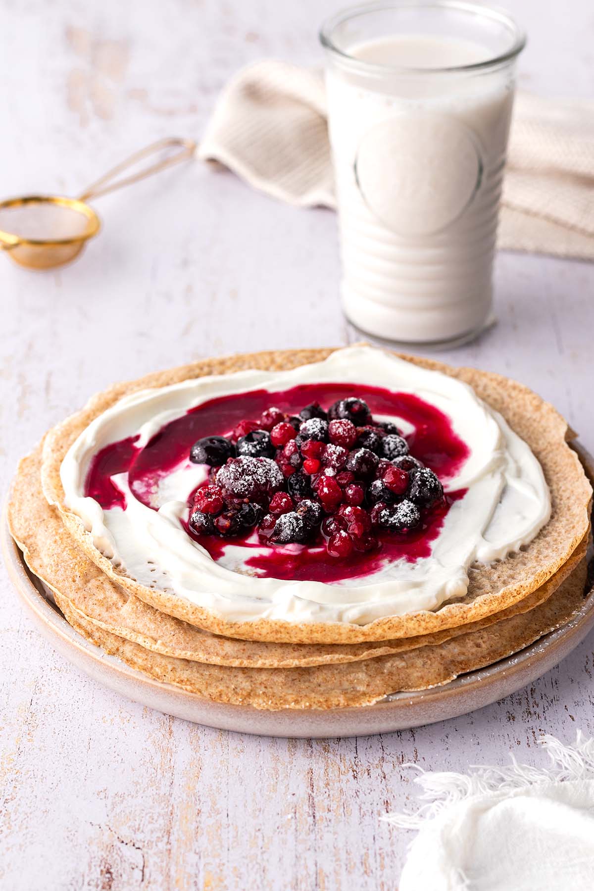 Ein Stapel Pfannkuchen auf einem Teller und einem Glas Milch im Hintergrund. Auf dem obersten Pfannkuchen ist Joghurt und hei&szlig;e Beeren.