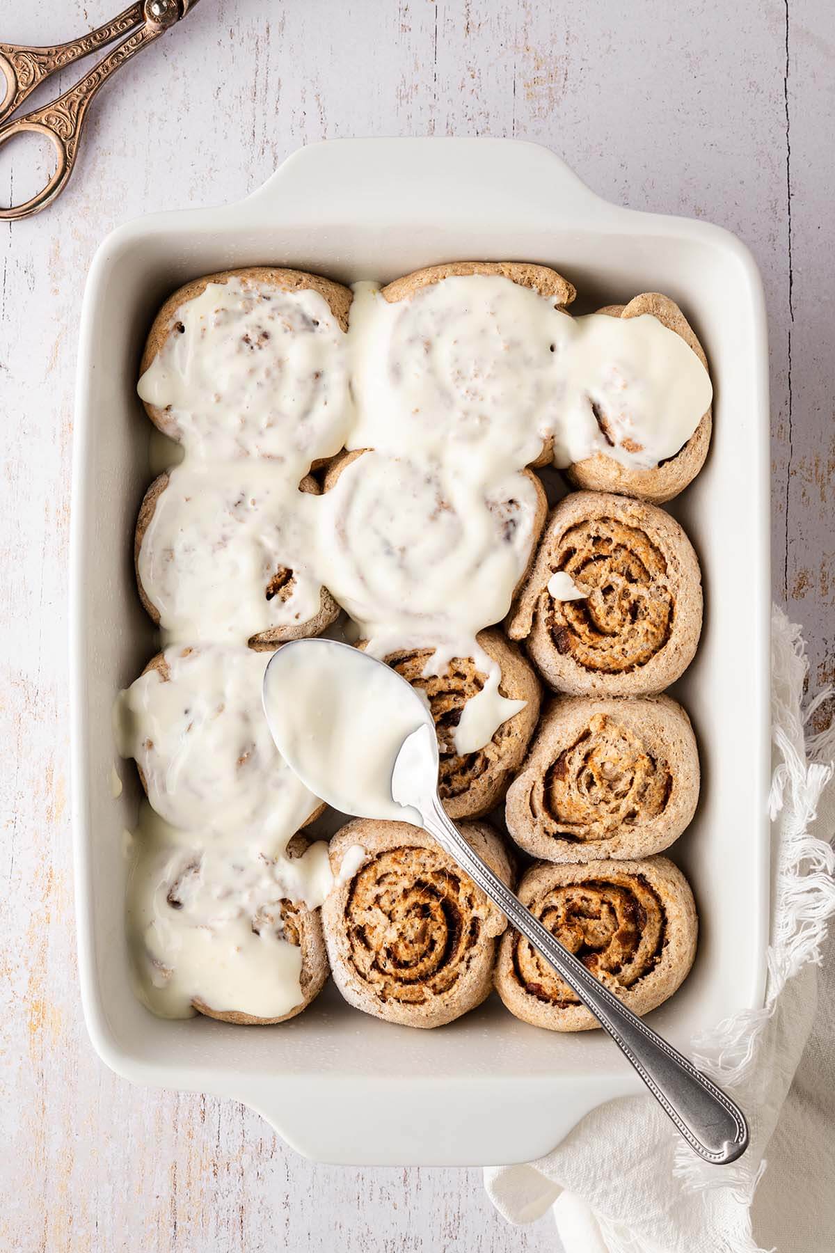 Mit einem L&ouml;ffel wird ein Frosting auf Zimtschnecken verteilt, die in einer Auflaufform sind.