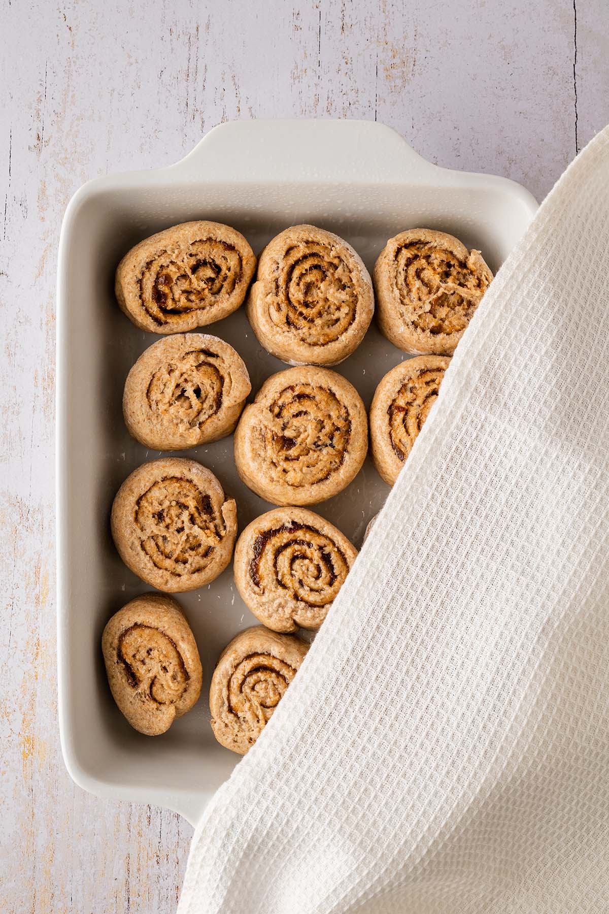 Eine halb mit einem Tuch abgedeckte Auflaufform mit rohen Zimtschnecken drin auf einem wei&szlig;en Holztisch.