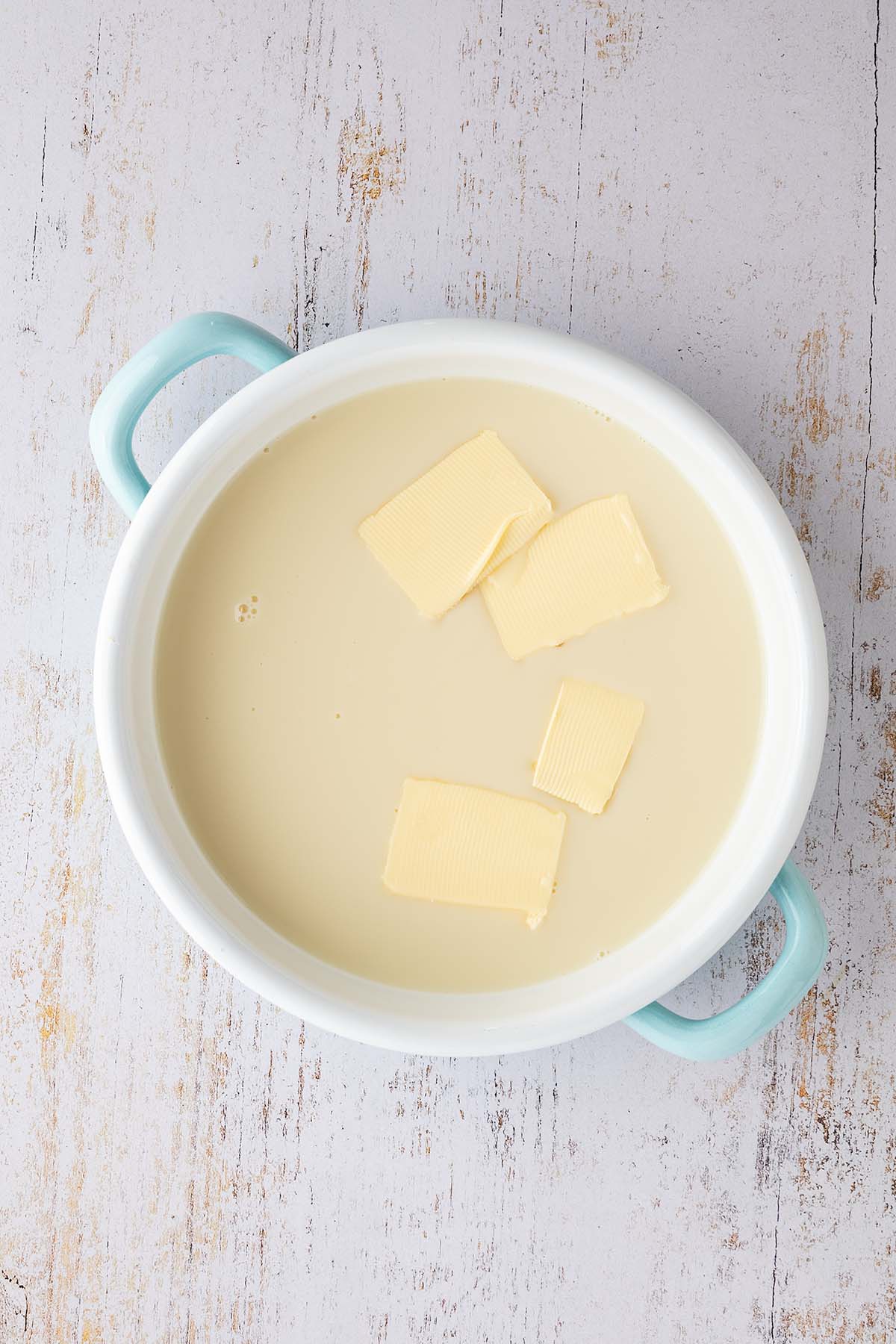 Eine kleine Emaille-Pfanne gef&uuml;llt mit Milch und Butter auf einem wei&szlig;en Holzuntergrund.
