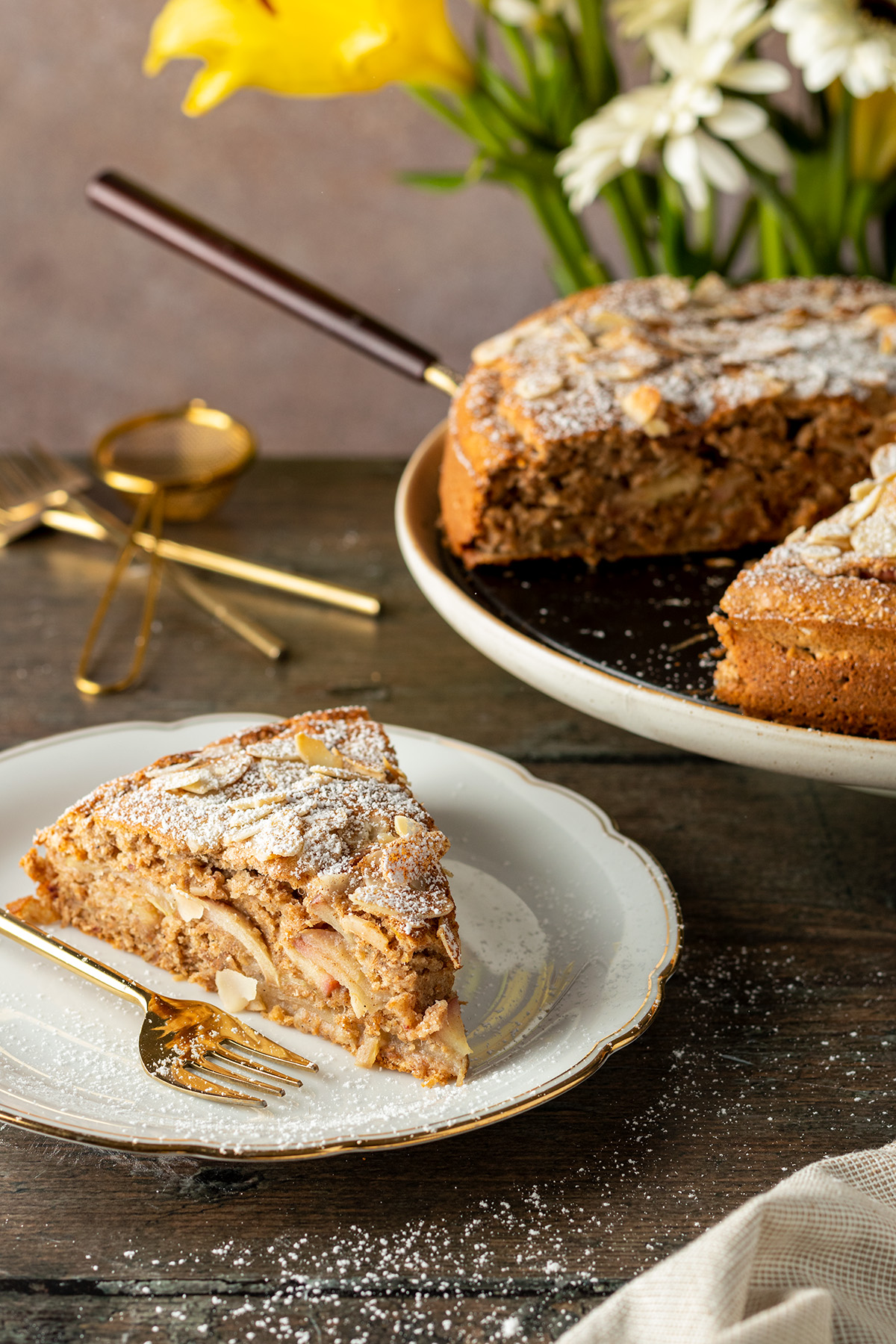 Ein Holztisch mit einem St&uuml;ck gesunden Apfelkuchen auf einem wei&szlig;en Teller und dem restlichen Kuchen im Hintergrund.