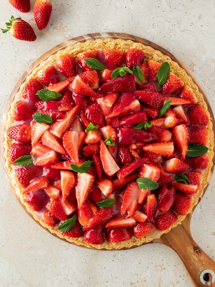 Gesunder Erdbeerkuchen mit frischen Erdbeeren und Minze auf einem runden Holzbrett, das auf einem steinernen Untergrund steht.
