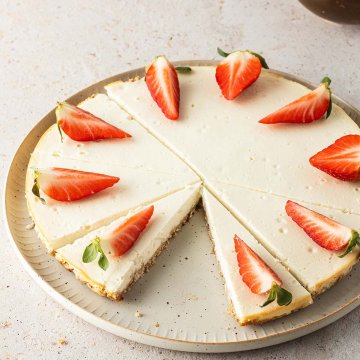 Ein K&auml;sekuchen serviert auf einem Teller mit frischen, geviertelten Erdbeeren, eine Schale Erdbeeren im Hintergrund.