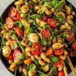 Bunter Nudelsalat mit Tomaten und Rucola auf einem gro&szlig;en Teller mit Deko im Bildrand.